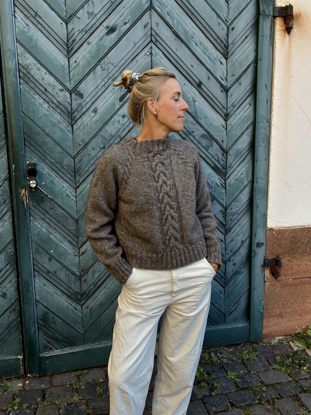 Frau im graubraunen Seestück Sweater mit großem Mittelzopf vor einem grünen Holztor
